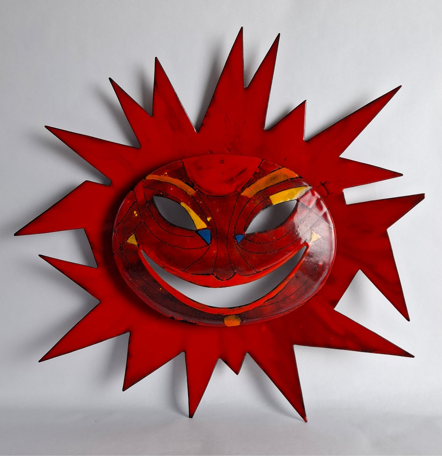 An enamel wall mask with a sunburst design featuring a smiling face in red, yellow, and blue enamel.