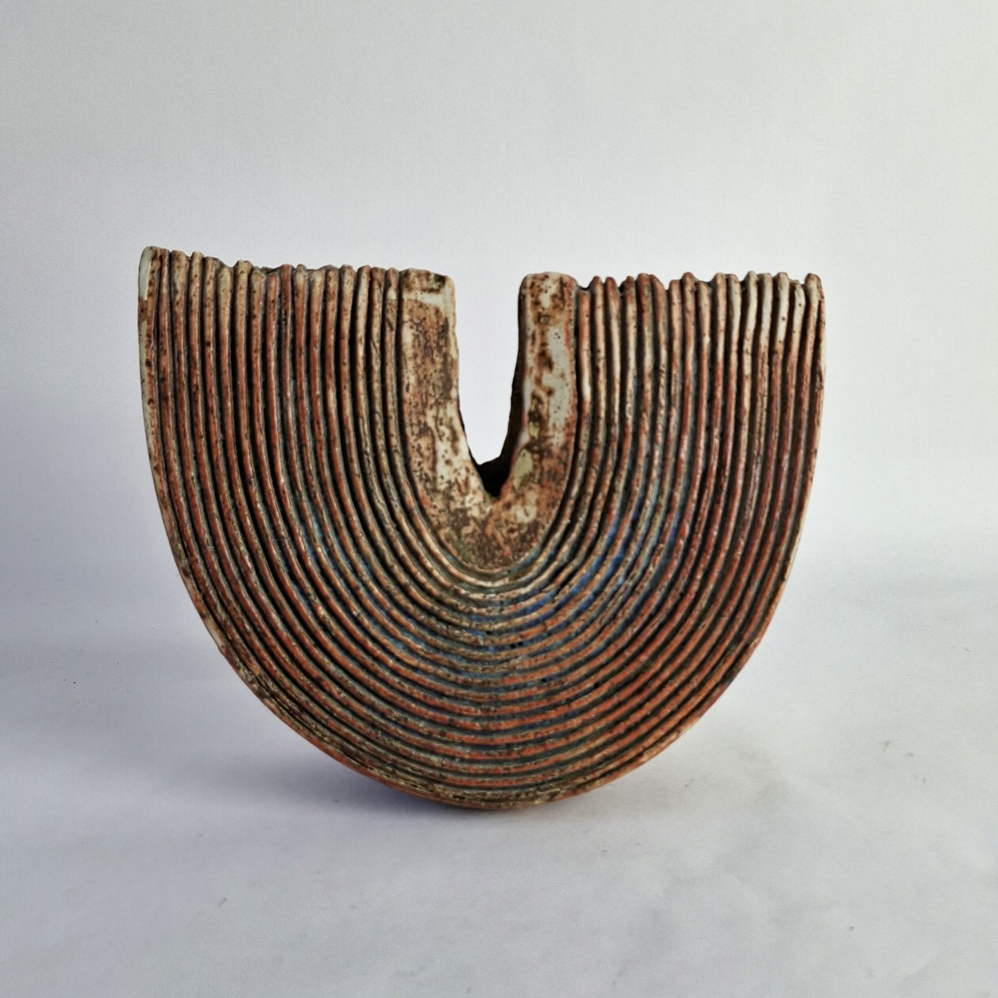 A grooved slice form stoneware vase with a deep rusty red glaze and accents of greens, blues, and turquoise.