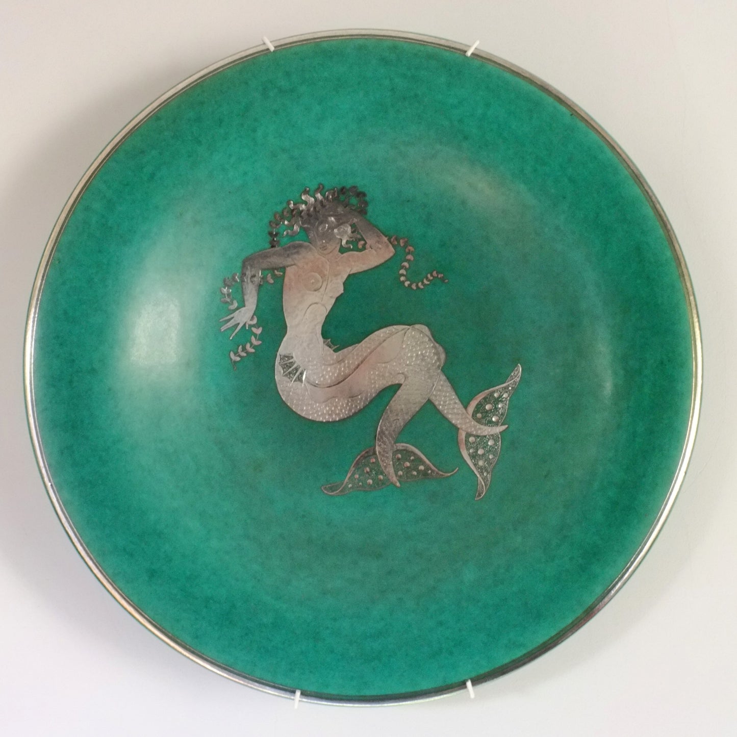 A stoneware charger with a central silver mermaid decoration on a green background, with a silver rim.