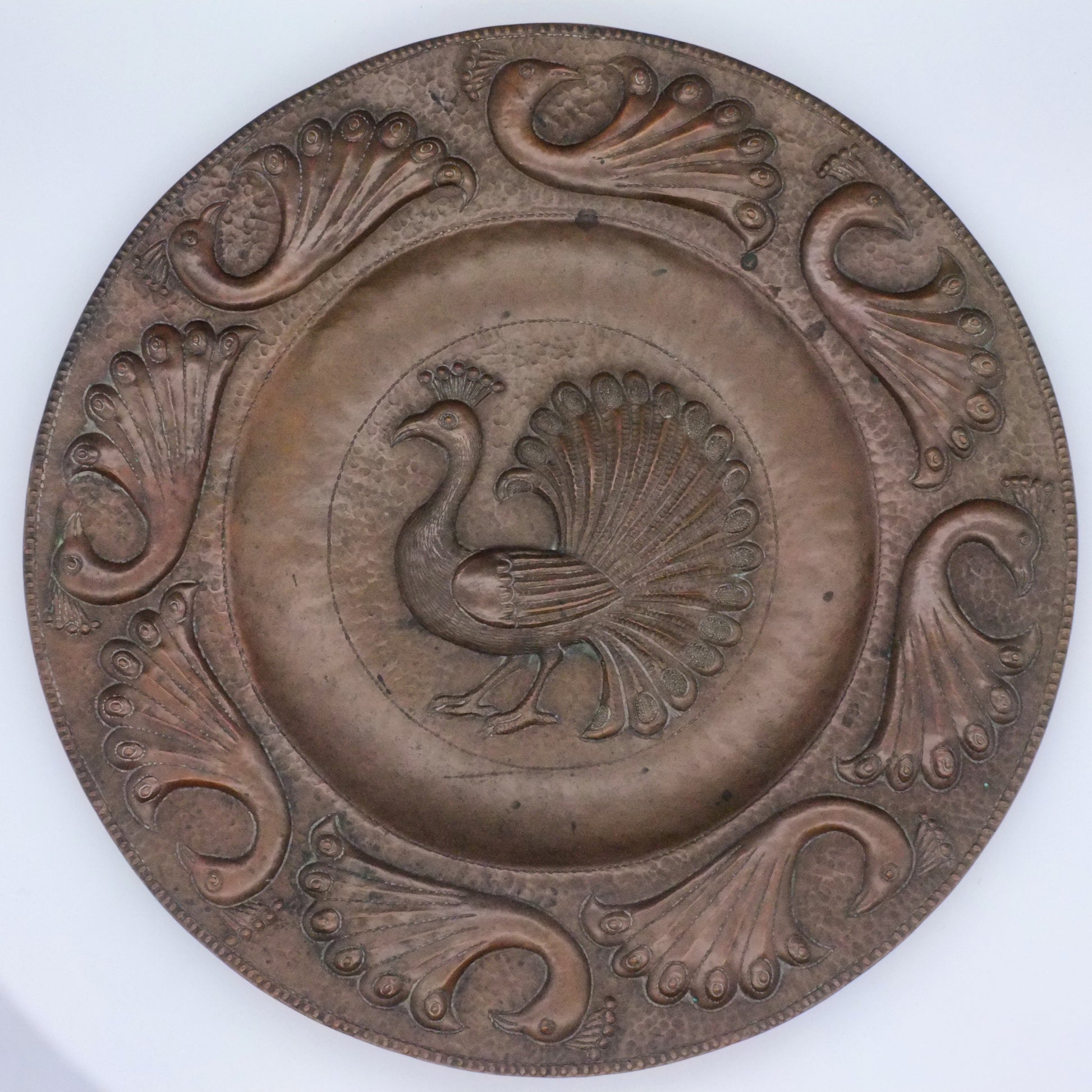 A copper charger with a central motif of a standing peacock, surrounded by eight stylized peacock designs around the edge.
