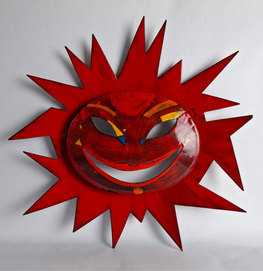 An enamel wall mask with a sunburst design featuring a smiling face in red, yellow, and blue enamel.
