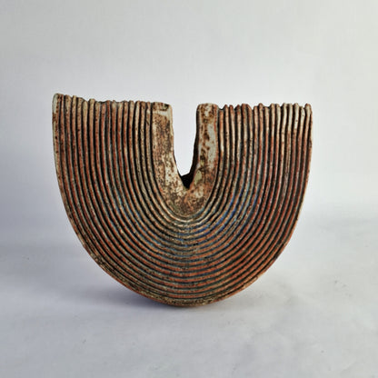 A grooved slice form stoneware vase with a deep rusty red glaze and accents of greens, blues, and turquoise.