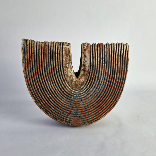 A grooved slice form stoneware vase with a deep rusty red glaze and accents of greens, blues, and turquoise.