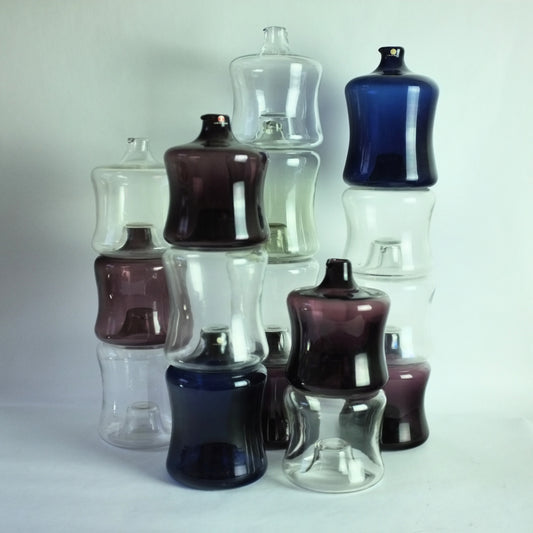 A collection of 16 stacking glass bottles designed by Timo Sarpaneva, featuring various colors such as clear, purple, blue, and pale green.